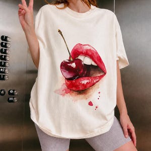 Op de afbeelding: Crèmekleurig T-shirt met een aquarelontwerp van rode lippen en een kers. De kers is gepositioneerd alsof erin wordt gebeten. Het shirt heeft een relaxte pasvorm en een ronde hals.