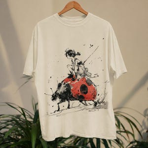 May include: White t-shirt featuring a black and red illustration of a person riding a ladybug. The artwork has a sketch-like style with splatters and fine lines. The ladybug is red with black spots, and the person is in a flowing outfit.