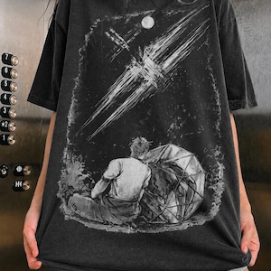 May include: A charcoal gray short-sleeve t-shirt featuring a graphic print of a person sitting on the ground next to a large geometric sphere. Above them, a bright celestial streak cuts across a dark sky with a small moon in the background.