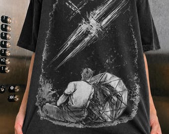 May include: A charcoal gray short-sleeve t-shirt featuring a graphic print of a person sitting on the ground next to a large geometric sphere. Above them, a bright celestial streak cuts across a dark sky with a small moon in the background.