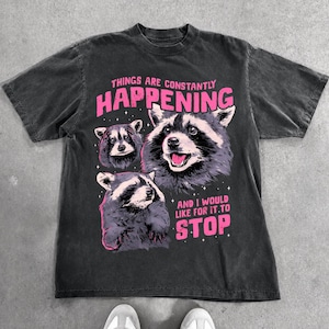 May include: A charcoal grey short-sleeve t-shirt featuring a graphic of three raccoons in shades of grey and black. The pink text reads: THINGS ARE CONSTANTLY HAPPENING AND I WOULD LIKE FOR IT TO STOP. The shirt has a crew neckline and a faded finish.