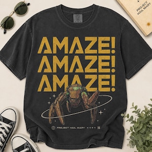 May include: Black t-shirt with the word "AMAZE!" repeated three times in gold. A brown and green insect-like creature is in the center, surrounded by a white ring. The shirt also features the text "PROJECT HAIL MARY".