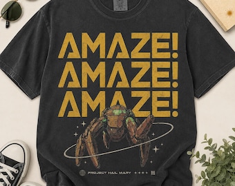 Könnte beinhalten: Schwarzes T-Shirt mit dem Wort "AMAZE!" dreimal in Gold wiederholt. Ein braunes und grünes insektenartiges Wesen befindet sich in der Mitte, umgeben von einem weißen Ring. Das Shirt hat auch den Text "PROJECT HAIL MARY".