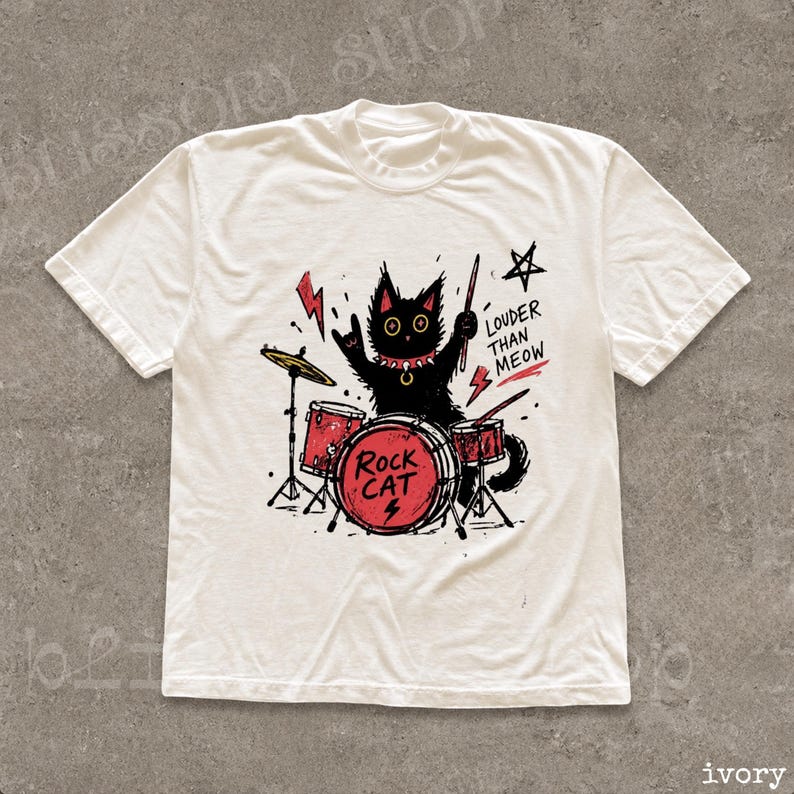 May include: An ivory short-sleeve t-shirt featuring a black cat playing red drums. The graphic includes a red lightning bolt, a black star, and the text "ROCK CAT" and "LOUDER THAN MEOW" in black lettering. The cat wears a spiked collar.