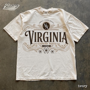 May include: An ivory short-sleeve t-shirt featuring a brown graphic print. The design includes the text "SMALL TOWN HEART", "VIRGINIA", "COUNTRY ROOTS", and "1788" surrounded by decorative scrollwork and floral accents. A circular logo with the letters "VA" is centred at the top.