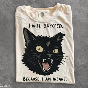 May include: An ivory t-shirt featuring a graphic of a black cat with yellow eyes and an open mouth showing teeth. Above the cat is the text I WILL SUCCEED, and below the cat is the text BECAUSE I AM INSANE. The cat has a small mark on its forehead.