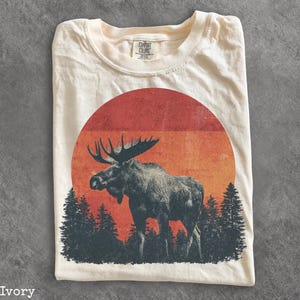 May include: An ivory t-shirt featuring a graphic print of a moose standing in front of a forest of pine trees and a large, circular, multi-toned orange and red sunset. The shirt has a Comfort Colors brand label printed on the inside neck area.