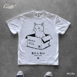 K&ouml;nnte beinhalten: Ein wei&szlig;es kurz&auml;rmeliges T-Shirt mit einer schwarzen Grafik einer Katze, die in einem Pappkarton sitzt. Der Karton tr&auml;gt den Text '拾われた rescued but still not paid' und das T-Shirt enth&auml;lt den Text '気にしない don't care' mit einem Pfotenabdruck.