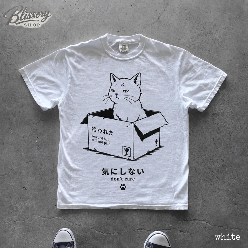 K&ouml;nnte beinhalten: Ein wei&szlig;es kurz&auml;rmeliges T-Shirt mit einer schwarzen Grafik einer Katze, die in einem Pappkarton sitzt. Der Karton tr&auml;gt den Text '拾われた rescued but still not paid' und das T-Shirt enth&auml;lt den Text '気にしない don't care' mit einem Pfotenabdruck.