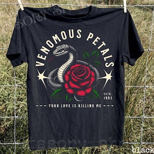 Peut inclure: Un t-shirt noir à manches courtes avec un motif représentant un serpent enroulé et une rose rouge. Le texte indique VENOMOUS PETALS, ESTD. 1993 et YOUR LOVE IS KILLING ME. Le design inclut deux étoiles à quatre branches et des feuilles vertes.
