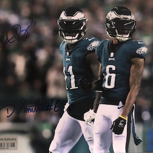 Puede incluir: Dos jugadores de fútbol americano con uniformes verdes y blancos, con cascos, están uno al lado del otro. La imagen está firmada con tinta morada. Los jugadores llevan guantes negros y pantalones blancos. El fondo está borroso.