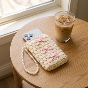 May include: A cream-colored crocheted phone pouch featuring three small light pink ribbon bows arranged vertically on the front. The pouch includes a white faux pearl beaded wrist strap and contains a smartphone with a visible camera array.