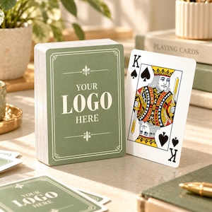 May include: A deck of playing cards with a sage green back featuring a white border, decorative flourishes, and the text YOUR LOGO HERE. Beside the deck stands a single King of Spades playing card with traditional red, yellow, and black colors.
