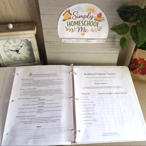 Puede incluir: Un archivador blanco abierto en una p&aacute;gina de "Reading Progress Tracker". El archivador est&aacute; sobre una superficie de color claro, con un reloj y un letrero decorativo que dice "Simply Homeschool Me" en el fondo. El rastreador tiene secciones para habilidades de lectura.