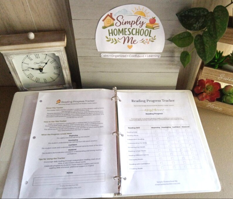 Puede incluir: Un archivador blanco abierto en una p&aacute;gina de "Reading Progress Tracker". El archivador est&aacute; sobre una superficie de color claro, con un reloj y un letrero decorativo que dice "Simply Homeschool Me" en el fondo. El rastreador tiene secciones para habilidades de lectura.
