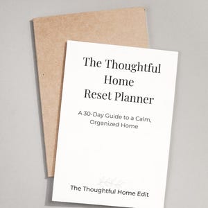 May include: A white planner titled "The Thoughtful Home Reset Planner" with the text "A 30-Day Guide to a Calm, Organized Home." The planner is on top of a brown envelope. The words "The Thoughtful Home Edit" are at the bottom.