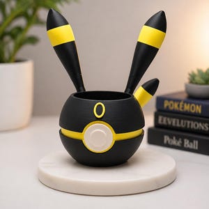 May include: A black and yellow Umbreon-themed desk organizer. The spherical container has two tall, black ears with yellow stripes, a yellow band, and a white circle with a yellow outline. It sits on a white marble coaster.
