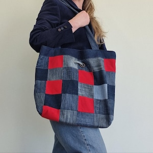 Puede incluir: Un bolso tote de mezclilla con un diseño de patchwork de cuadrados azules y rojos. El bolso tiene un cierre de cordón y una correa larga. Hecho de mezclilla reciclada, un accesorio único y ecológico.