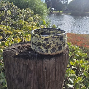 May include: A round ceramic bowl with a textured, cratered surface featuring a high-contrast color scheme of deep black and vibrant lime green. The bowl has a thick, uneven rim and a porous, rocky appearance throughout its exterior wall.