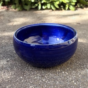 May include: A round, deep blue ceramic bowl with a glossy finish. The bowl has a simple, minimalist design and is sitting on a textured surface. The bowl's interior reflects the surrounding environment.