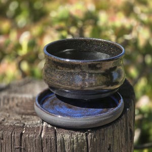 Könnte beinhalten: Ein handgefertigtes Keramikschalen- und Untertassen-Set. Die Schale ist dunkel mit metallischem Glanz, und die Untertasse hat eine tiefblaue Innenseite. Das Set wird auf einer verwitterten Holzoberfläche mit verschwommenem Naturhintergrund präsentiert.