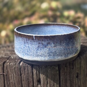 May include: A handmade ceramic bowl with a blue and brown glaze. The bowl has a round shape and a textured surface. The exterior is a dark brown, transitioning to a light blue interior. The bowl is sitting on a weathered wooden post.