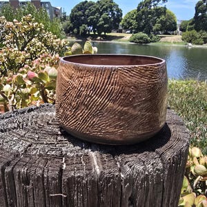 May include: A cylindrical ceramic bowl with a rich brown glaze and a textured, cross-hatched surface pattern. The bowl features a wide, open top and a smooth, dark interior, showcasing a handcrafted appearance with visible clay work details.