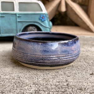May include: A handmade ceramic bowl with a blue and purple glaze. The bowl has a textured, horizontal striped pattern around the middle and a cream-colored base. A blue van is in the background.