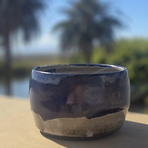 May include: A handmade ceramic bowl with a rounded shape. The bowl features a dark blue glaze on the upper portion, transitioning to a lighter, earthy tone towards the base. The bowl is sitting on a light-colored surface, with a blurred outdoor background.