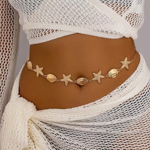 Puede incluir: Una cadena de cuerpo dorada con colgantes de conchas marinas y estrellas de mar. La cadena se lleva alrededor de la cintura. La modelo lleva un top y una falda blancos de tejido abierto. Accesorio de temática playera.