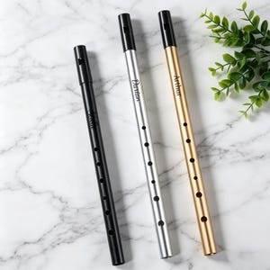 May include: Three tin whistles in black, silver, and gold, each with a black mouthpiece and engraved name. The black whistle has the name "Stella", the silver one "Dartan", and the gold one "Arthur". The instruments are on a marble surface.