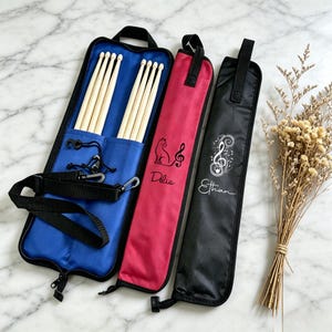 May include: Three drumstick cases in blue, red, and black. The blue case is open, displaying wooden drumsticks. The red case has a cat and musical note design with the name "Délia". The black case has a musical note design with the name "Ethan".