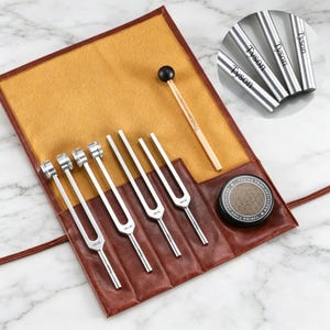 May include: A collection of silver tuning forks, each engraved with "Tyson", presented in a brown leather case with a wooden mallet. The case features a tan interior and a small, round container with text.