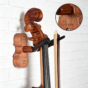 May include: A wooden violin wall mount with a violin and bow. The mount is a dark brown colour and has the name "Darien" engraved on it. The violin is brown and black, and the bow is light brown.