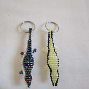 May include: Two beaded lizard keychains. One is primarily white with iridescent blue, green, and purple beads, and red eyes. The other is light green with black edging. Both have silver key rings.