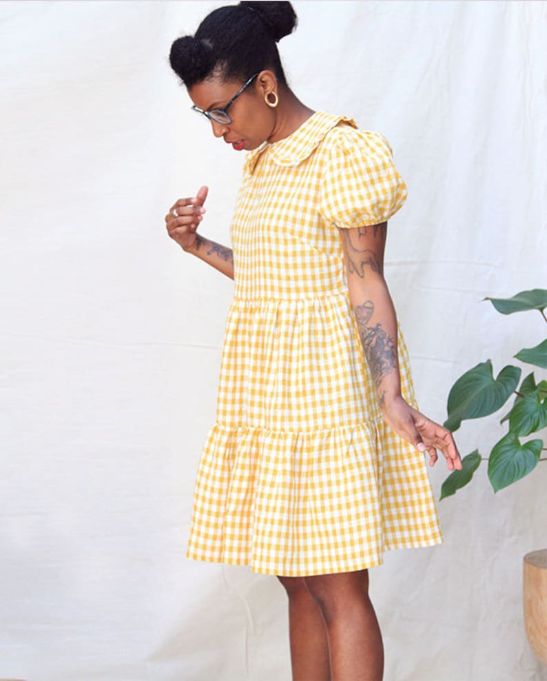 K&ouml;nnte beinhalten: Ein gelbes und wei&szlig;es Gingham-Kleid mit Peter-Pan-Kragen und Puff&auml;rmeln. Das Kleid hat einen Stufenrock und ist knielang.