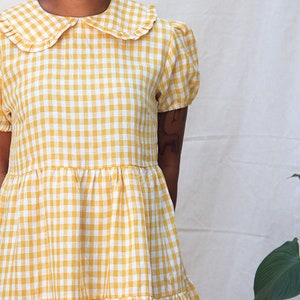 K&ouml;nnte beinhalten: Ein gelbes und wei&szlig;es Gingham-Kleid mit einem R&uuml;schenkragen und kurzen &Auml;rmeln. Das Kleid hat einen Stufenrock.