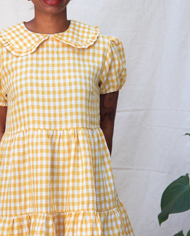 K&ouml;nnte beinhalten: Ein gelbes und wei&szlig;es Gingham-Kleid mit einem R&uuml;schenkragen und kurzen &Auml;rmeln. Das Kleid hat einen Stufenrock.