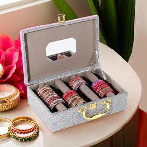 Puede incluir: Una caja de joyas brillante, con un asa y cierre dorados, llena de pulseras coloridas. La caja está forrada con tela gris y tiene un pequeño espejo en el interior. La caja está sobre una superficie blanca.