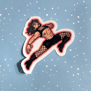 May include: A die-cut sticker featuring a stylized illustration of a person with long dark hair, wearing a black harness, black shorts, fishnet stockings, and black boots. The figure is posed in a dynamic, mid-air leap against a light blue background.