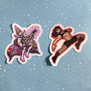 May include: Two die-cut stickers featuring illustrated figures. The left sticker shows a person in a rainbow-tipped cone hat and a colorful patchwork cape holding a large, crystalline, musical-note-adorned object. The right sticker depicts a person in a black harness, fishnet stockings, and black boots in a dynamic, leaping pose.