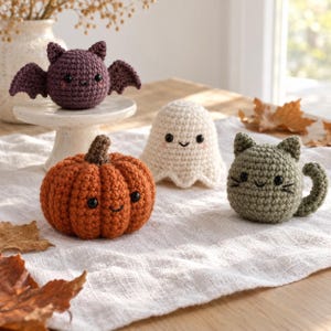 May include: A set of four crocheted figures including a purple bat on a white pedestal, an orange pumpkin, a white ghost, and a sage green cat. Each figure features black plastic eyes and embroidered facial details, crafted with yarn.