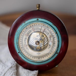 May include: A vintage barometer with a dark red wooden frame and a turquoise-colored ring. The face displays weather indicators like "Rain" and "Fair." A brass knob sits atop the circular instrument. The barometer is a classic weather forecasting tool.