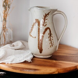Peut inclure: Une cruche en céramique crème avec un motif botanique marron. La cruche a une anse et un bec verseur. Elle repose sur une planche de service en bois avec un tissu blanc. Une bouteille en verre transparent avec des fleurs séchées est à l'arrière-plan.