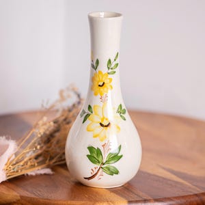 May include: A white ceramic vase with a bulbous base and a slender neck. The vase is adorned with hand-painted yellow flowers and green leaves. The vase is displayed on a wooden tray.