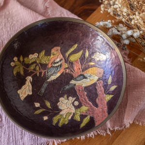 May include: A circular, dark purple bowl with a brass rim. The bowl has a painted design of two birds on a tree branch with leaves and flowers. The birds have orange, yellow, and blue feathers.