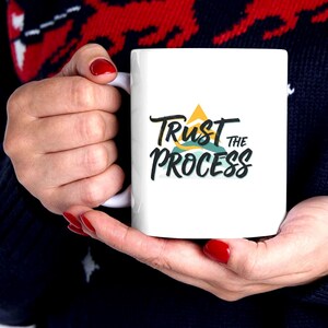 May include: White ceramic mug with the text "Trust the Process" in black script, with a yellow mountain and blue water graphic. The mug is held by a person with red nail polish, against a dark blue sweater with a red and white pattern.