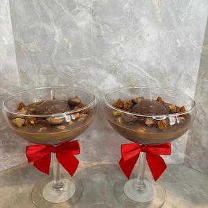 以下が含まれることがあります： 脚付きグラスに入った2つの装飾用キャンドル。キャンドルはチョコレートのような外観の茶色で、ナッツとチョコレートがトッピングされています。各グラスのステムには赤いリボンが付いています。
