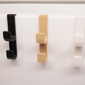 Op de afbeelding: Drie over-deurhaken in zwart, beige en wit. Elke haak heeft een geribbeld ontwerp en twee onderste haken om items op te hangen. De haken zijn gemaakt van een duurzaam materiaal en passen over een standaarddeur.