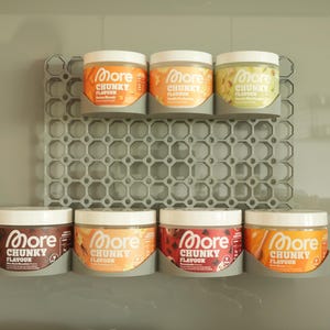 Könnte beinhalten: Sieben runde Kunststoffdosen mit More Chunky Flavour Würzpulver, angeordnet in einem grauen Wandregal mit Wabenmuster. Die Dosen haben weiße Deckel und verschiedenfarbige Etiketten mit aufgedrucktem Text in verschiedenen Farben.